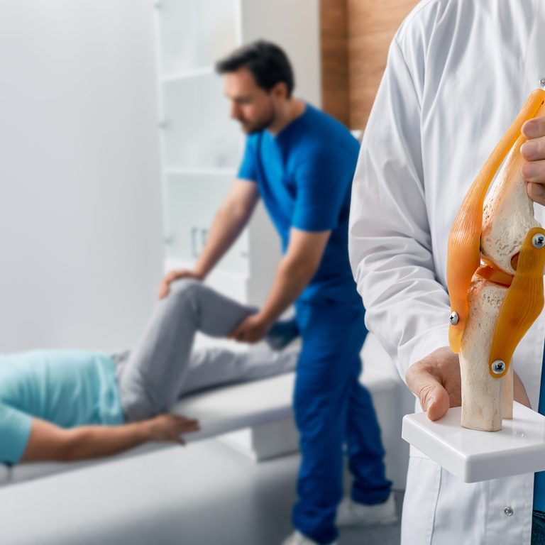 Physiotherapist holding anatomical human knee-joint model in hands, over background physiotherapy session for male patient. Physiotherapy treatments and kinesiotherapy, conceptual image